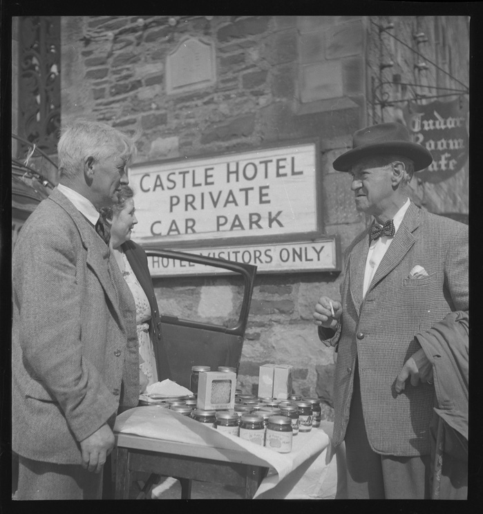 A photograph from 1950 shows two men and a woman engaging in conversation at the Conwy Honey Fair near a table displaying jars, with a sign for 'Castle Hotel Private Car Park' in the background.